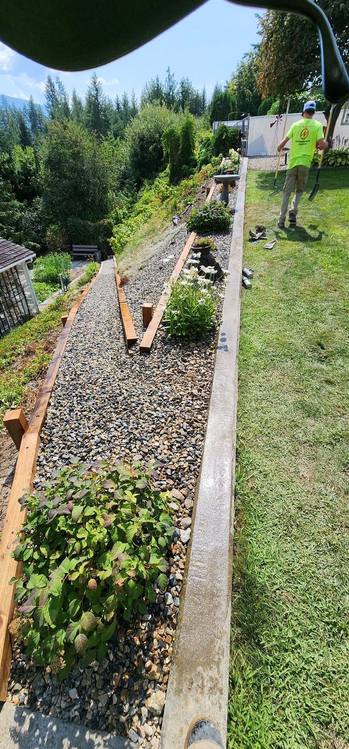 A person mows a lawn next to a terraced garden bed with gravel and plants, set against a hillside.