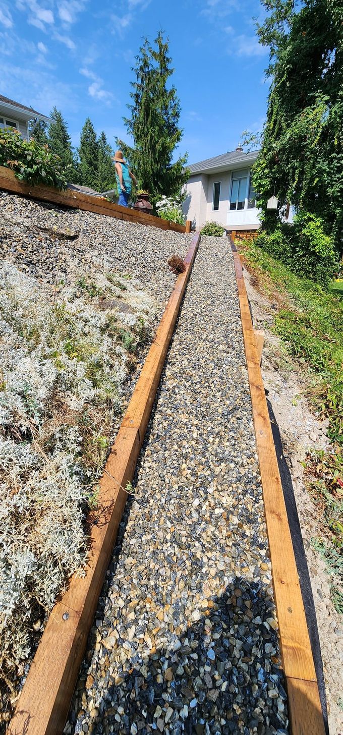 Gravel walkway bordered by wooden beams on a hillside, leading towards a house under a blue sky.