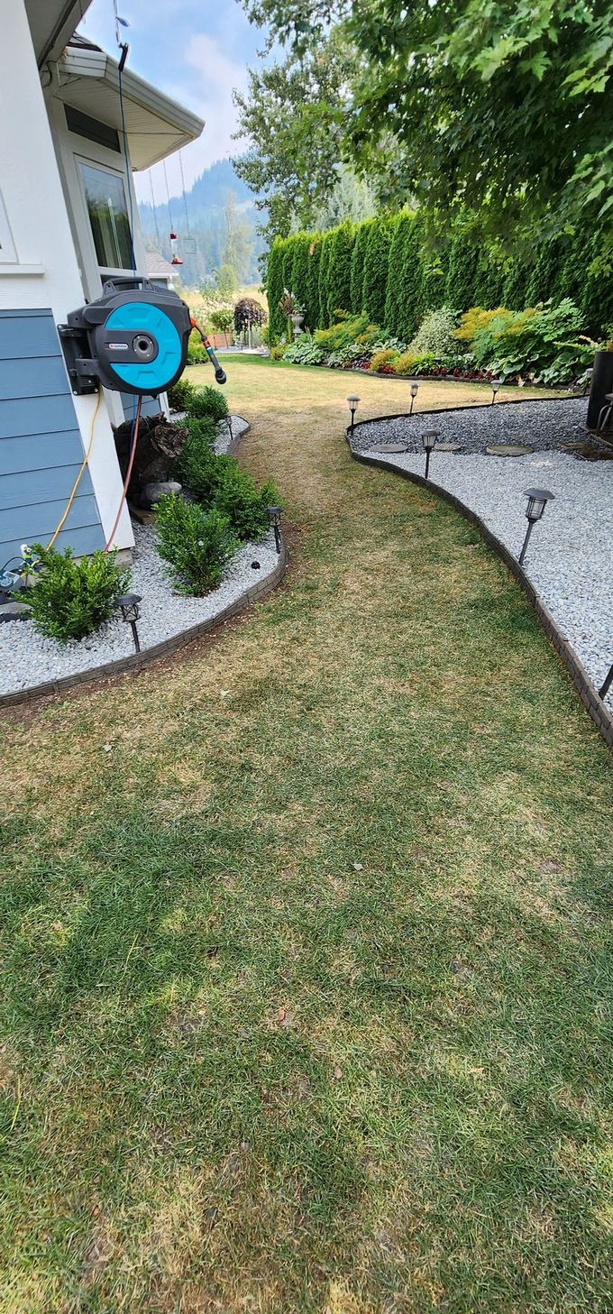 A garden path with a house on the left and a lush green area on the right, with a hose reel on the house.