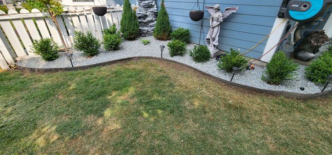 A well-manicured garden bed with various green plants, white pebbles, and a brown border. Green lawn in the foreground.