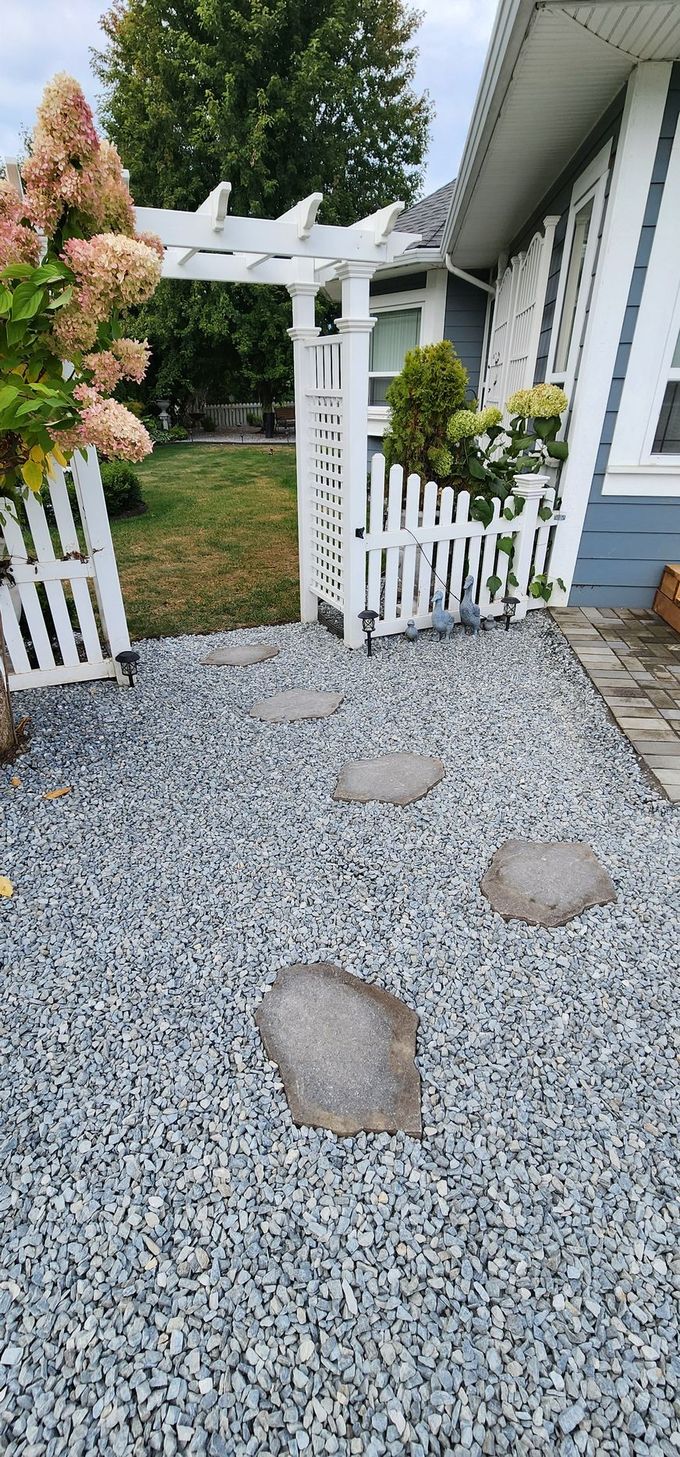 Gray gravel pathway with stepping stones leads to a white picket fence and trellis. Pink flowers and green trees in background.