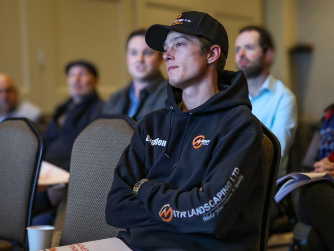 Young man in black hoodie and cap, arms crossed, seated at a conference; looking off to the side.