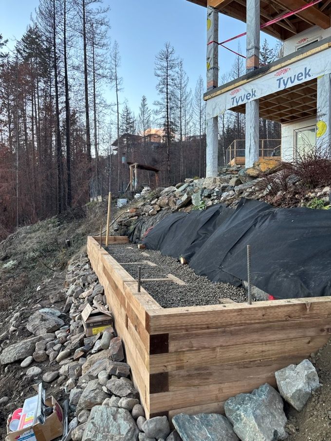 Wooden retaining wall construction on a hillside, with a house in the background and rocky terrain.