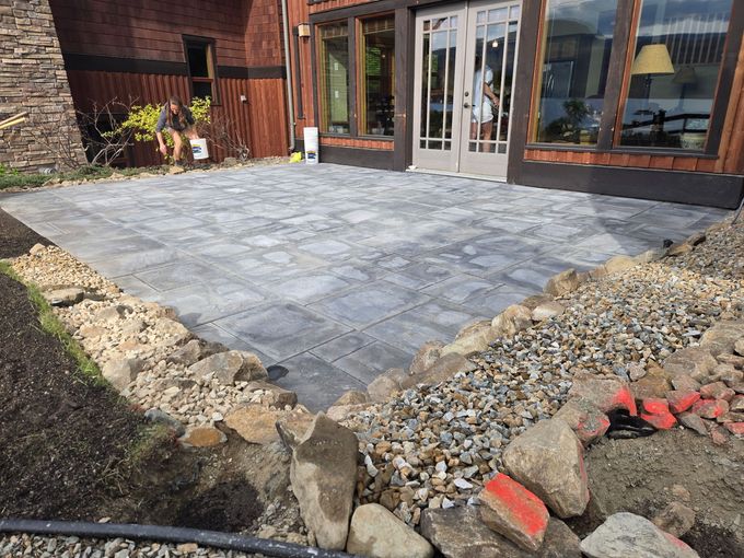 Patio under construction with gray pavers, dirt, rocks, and a building. A person is working nearby.