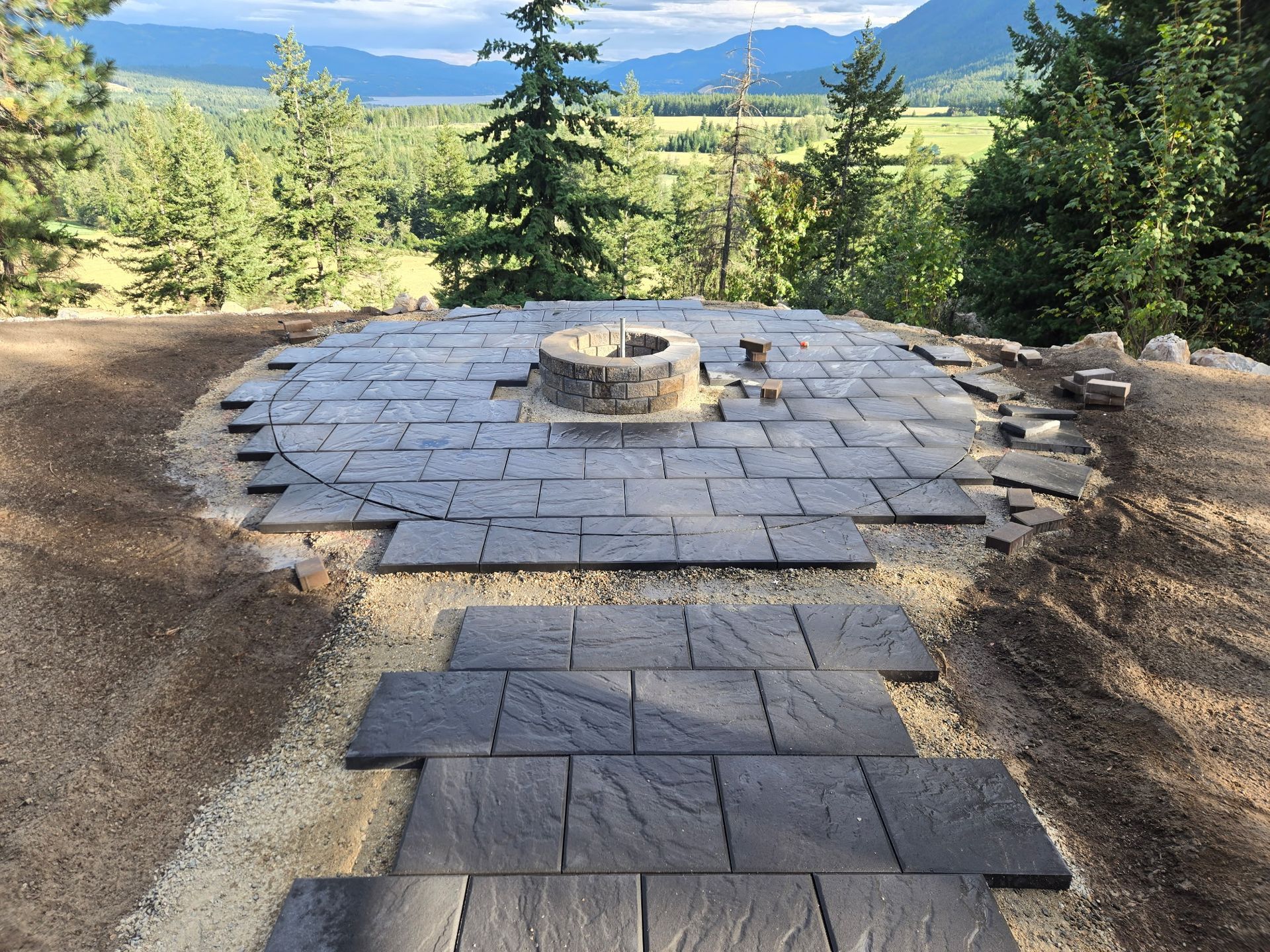A partially completed patio with steps, a fire pit, and a scenic mountain and forest background.