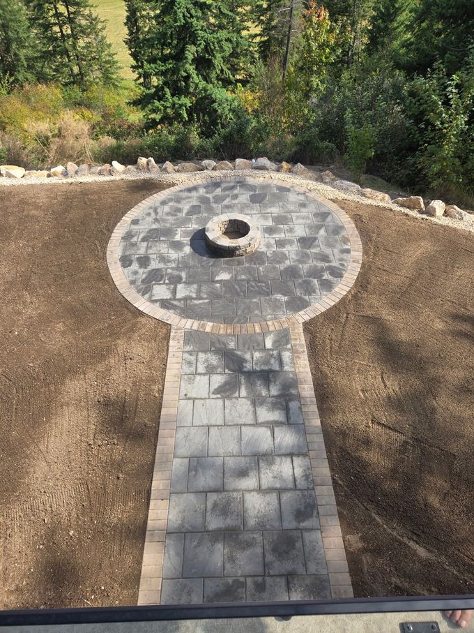 Overhead view of a circular patio with a fire pit, connected by a pathway, surrounded by dirt and trees.
