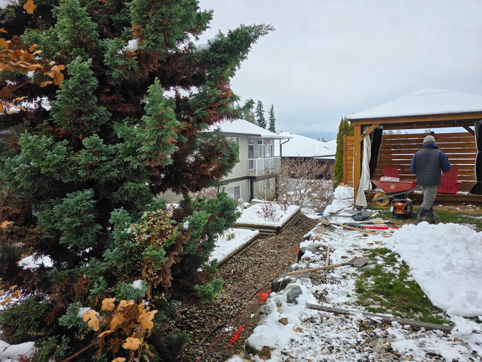 Snowy backyard scene with a person near a wooden gazebo, a large tree on the left, and a house in the background.