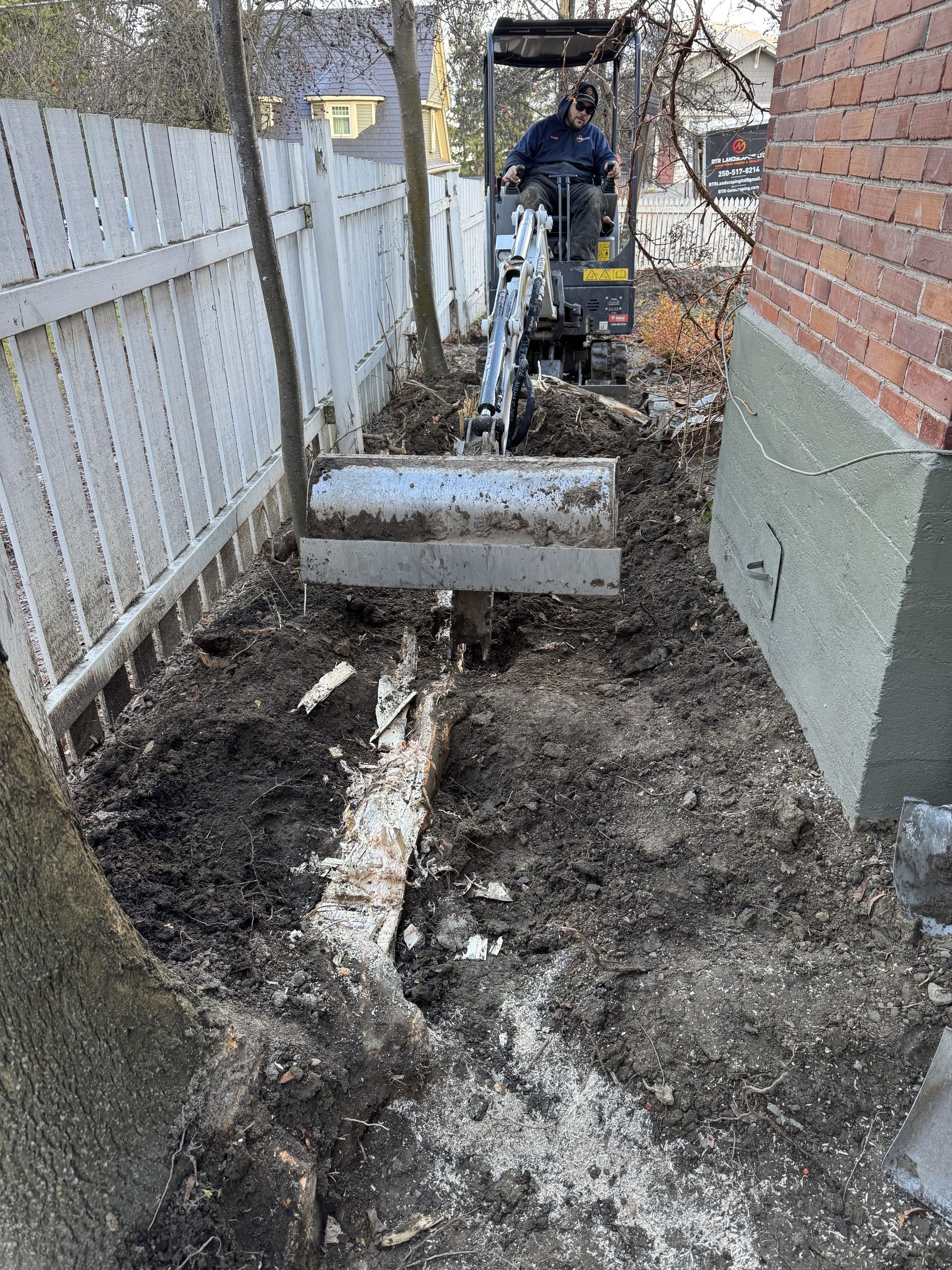 A person operating a small excavator digs near a white fence and a brick building.