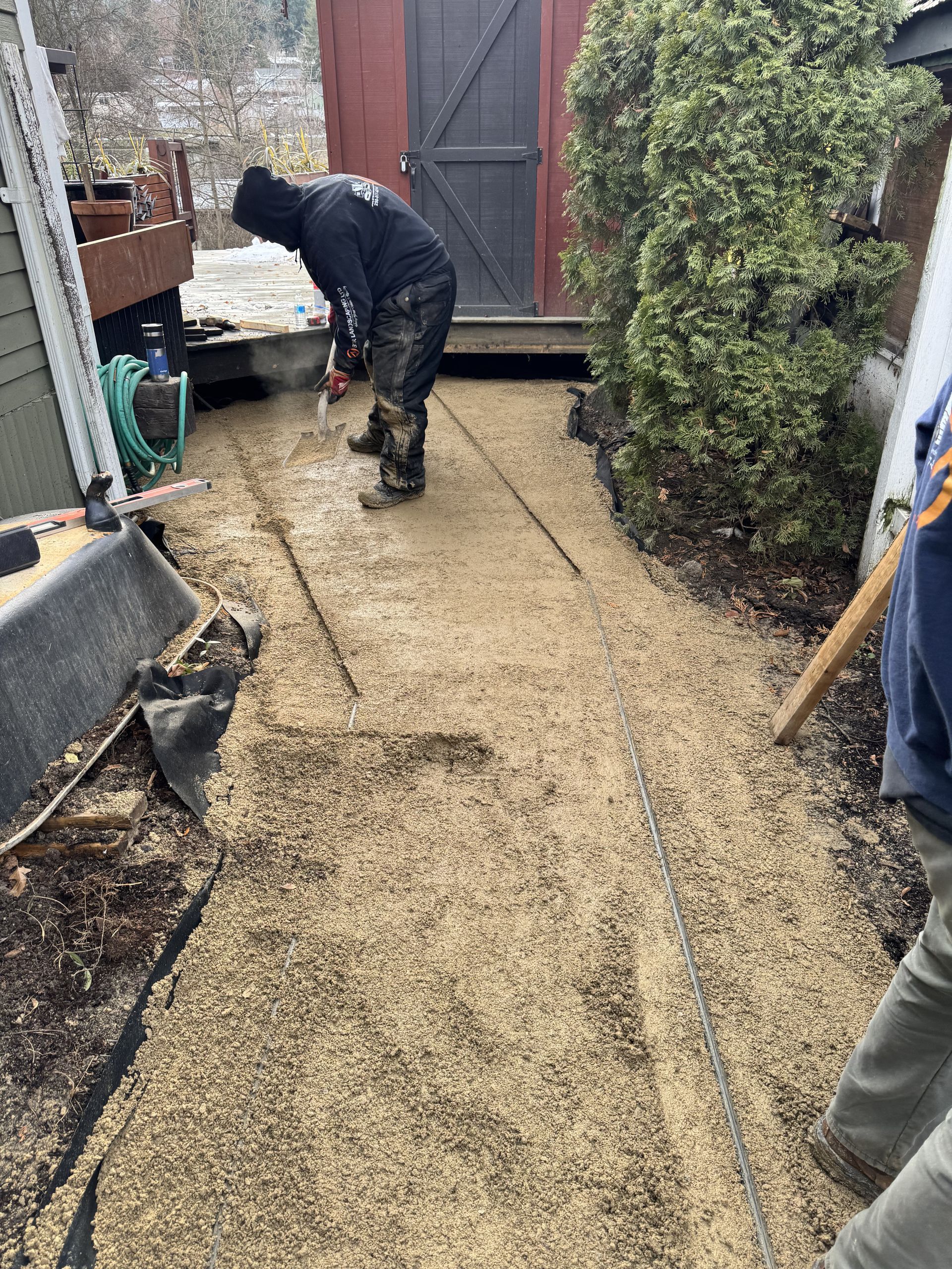 Person spreading gravel on a walkway. Setting is outdoors with a shed and bush in the background.