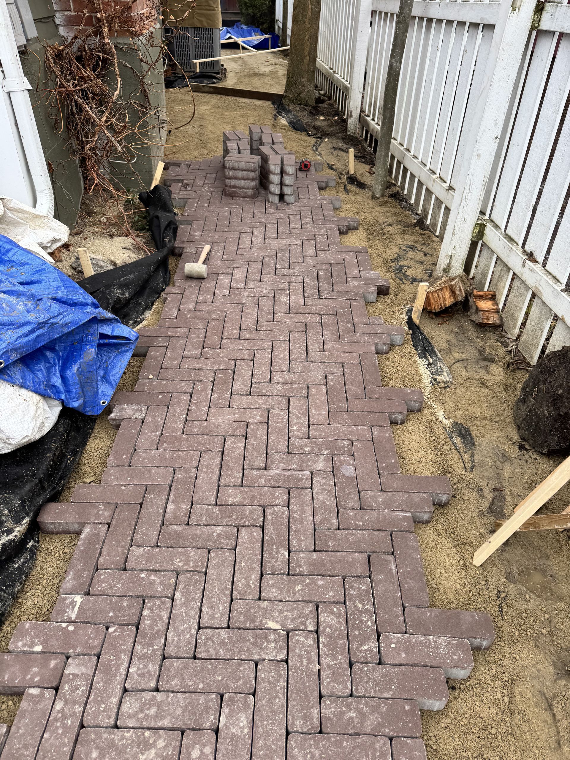 Brick pathway under construction; herringbone pattern, red-brown bricks on sand base, next to a white fence.