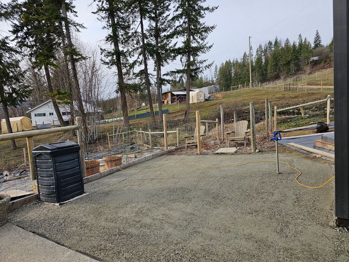 A gravel patio with a compost bin, fence, and view of trees, hillside, and a distant house.
