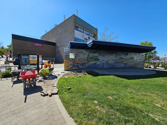 Workers near a colorful mosaic stage, park setting. A brick building and a storefront in the background on a sunny day.