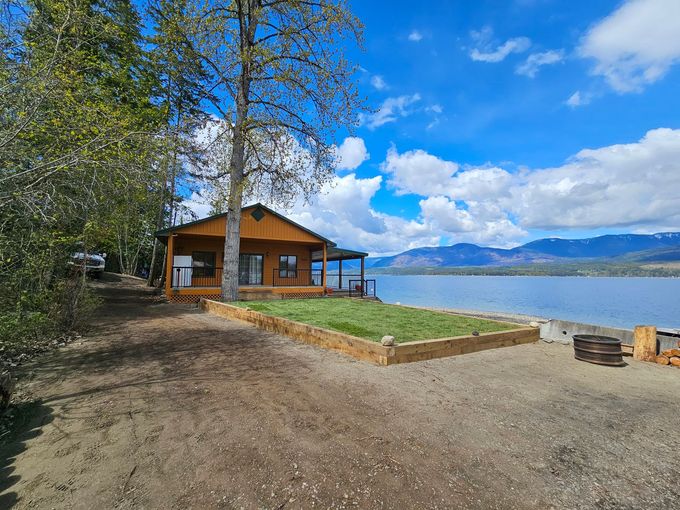 Lakeside cabin on a sunny day; brown wood, blue water, green grass, mountains in the distance.