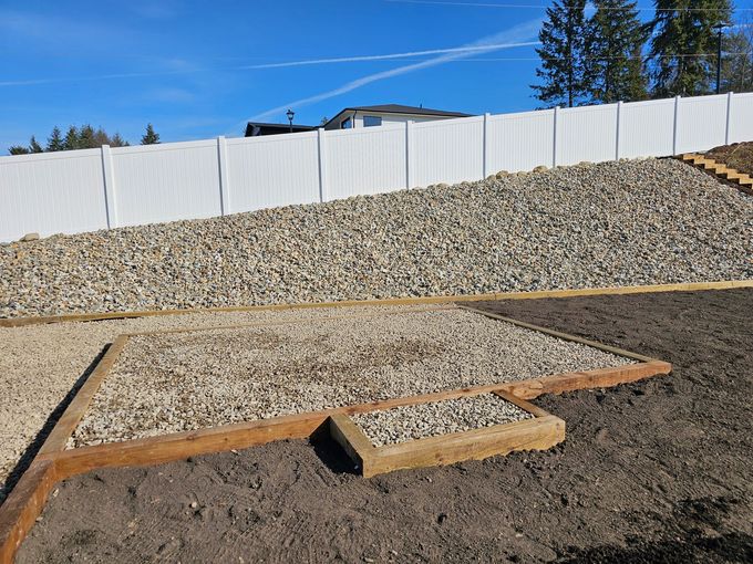 A gravel-covered hillside with a white fence. A wooden-framed area with gravel sits in the foreground.