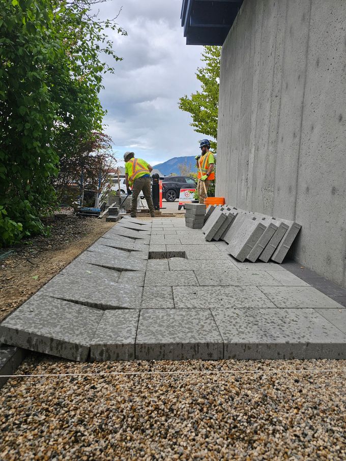 Construction workers laying gray paving stones next to a building; gravel bed, trees, cloudy sky.