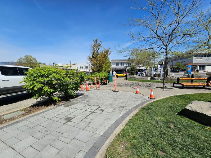 Brick walkway with orange cones and a small tree, leading to a yellow vehicle, park benches, and buildings under a blue sky.