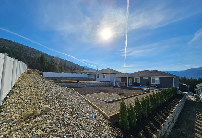 Sunny landscape with a house and retaining walls, gravel, and trees under a bright blue sky.