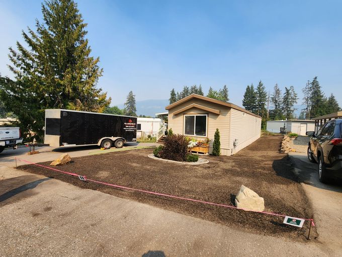A tan mobile home with fresh mulch landscaping, a black trailer, and a sunny outdoor setting.
