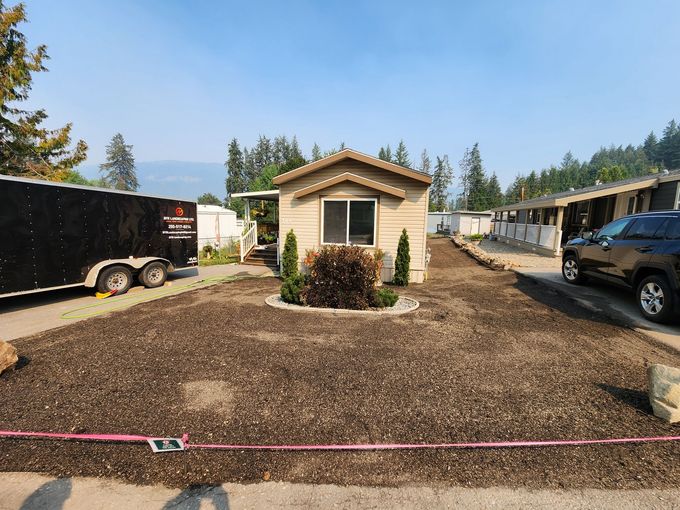 Mobile home exterior with landscaping; dark brown mulch, small trees, trailer and car parked in front.