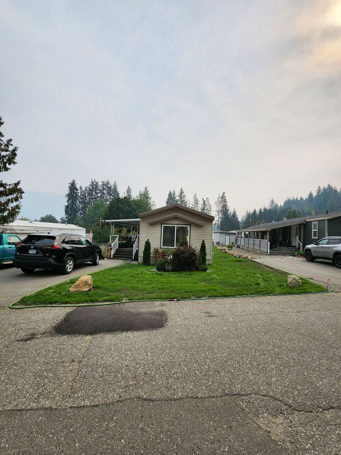 A small wood-paneled shed with a window sits on a patch of green grass, surrounded by cars and a parking lot.