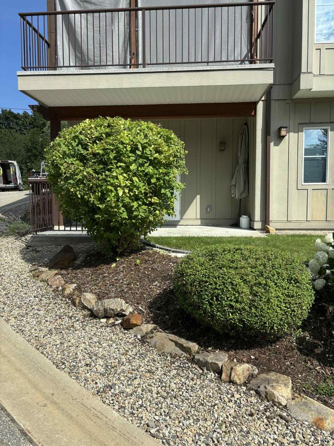 Exterior of a two-story building with a balcony. Two green bushes are in front of the building, with a gravel pathway in front.