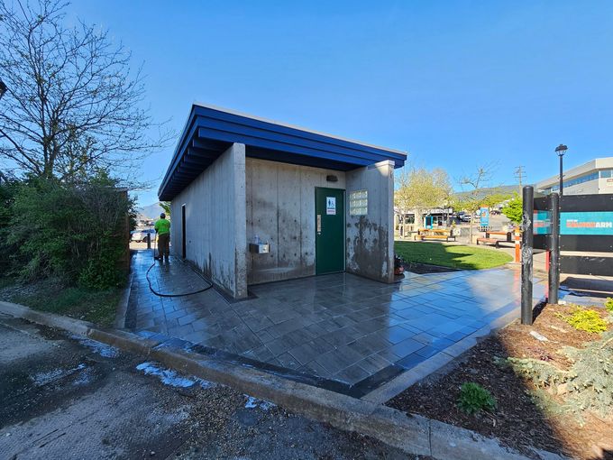A man power washes a concrete public restroom with a blue roof on a sunny day.