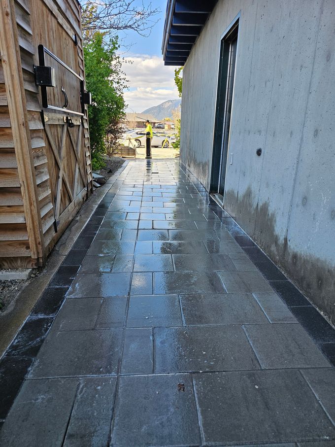 Brick walkway between wooden fence and building. Wet surface reflects light. Person in distance.
