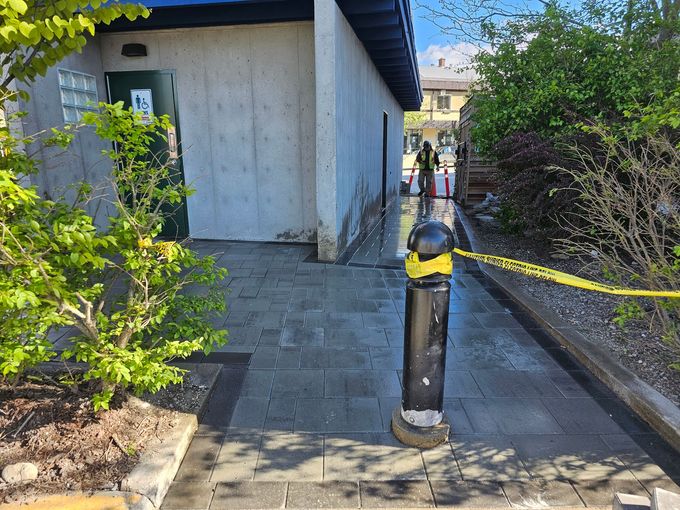 Sidewalk with a black bollard, caution tape, and a concrete building on the left. Green bushes are in the foreground.