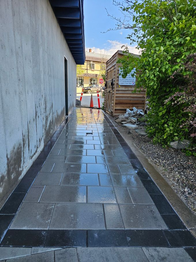 Wet brick walkway between a white wall and greenery, leading toward buildings.