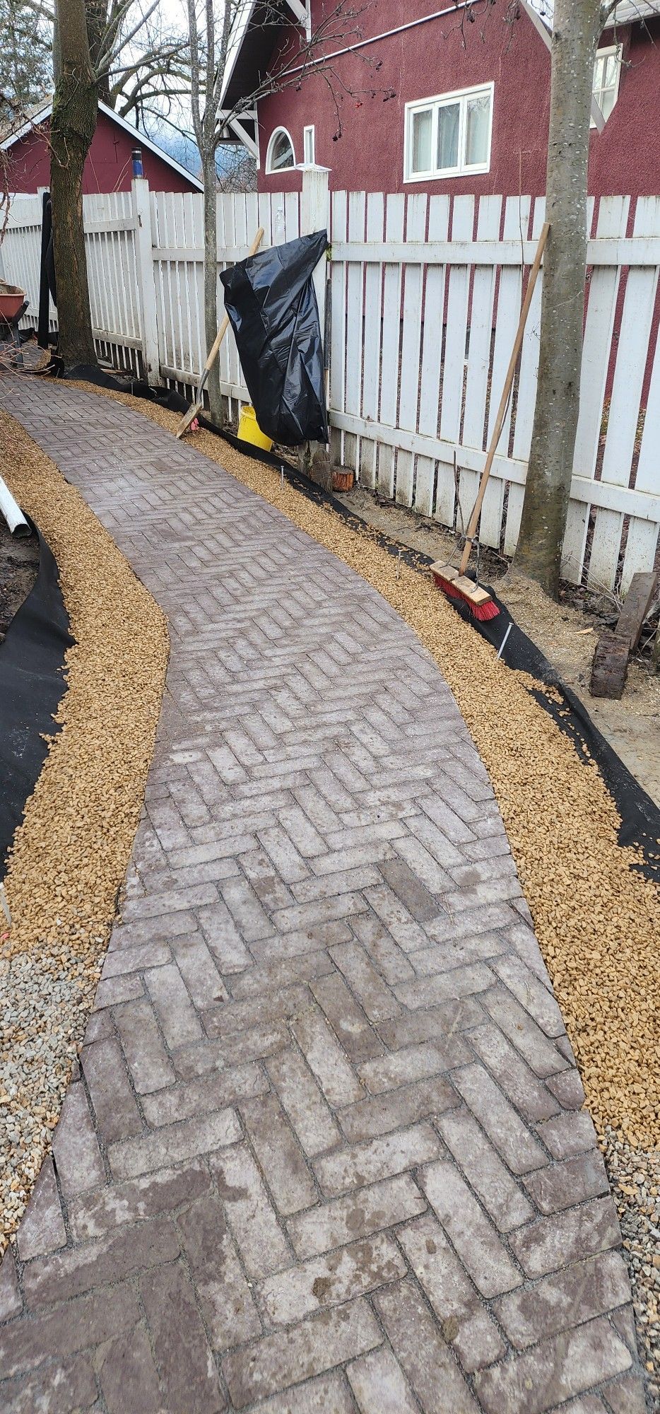 A brick pathway winding alongside a white fence and a red building, with gravel borders and trees.