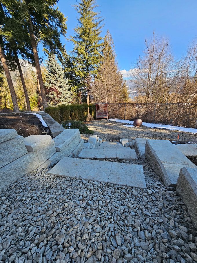 Gravel and stone steps in a yard with trees and patches of snow under a blue sky.