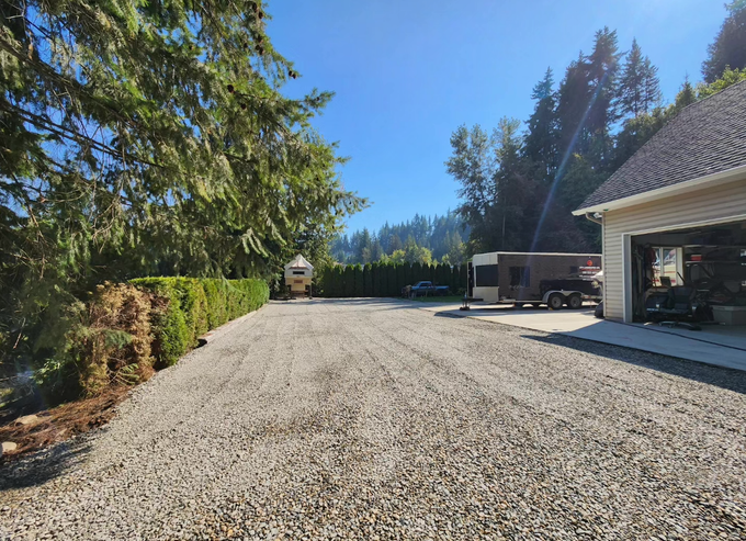 Gravel driveway in front of a house with a garage, trees, and a blue sky.