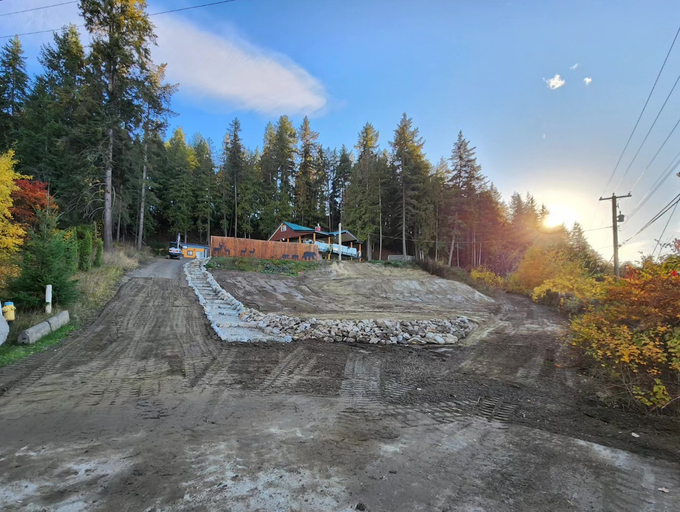 Construction site on a hillside: dirt, rocks, retaining wall. House visible in background, trees, blue sky, and setting sun.