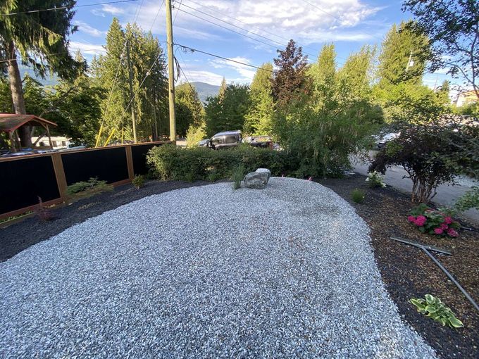 Gravel landscaping bed with a stone, bushes, and trees under a blue sky.
