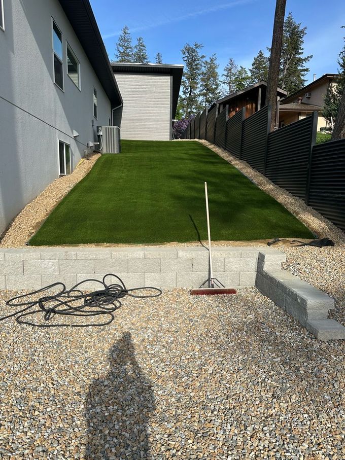 A backyard with a raised lawn, gravel, and a retaining wall. There's a hose and rake.