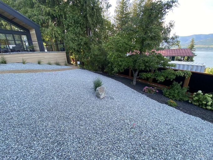 Gravel yard sloping toward a body of water, with a modern house on the left and trees on the right.
