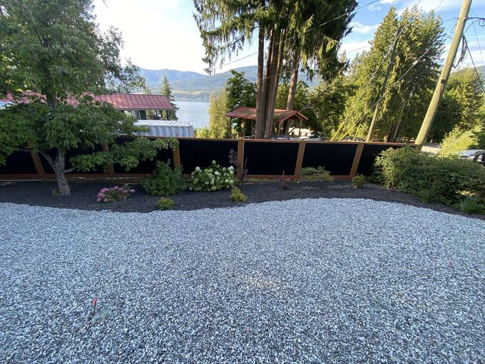 Gravel driveway with a black fence bordering a flowerbed, trees, and a lake in the background.