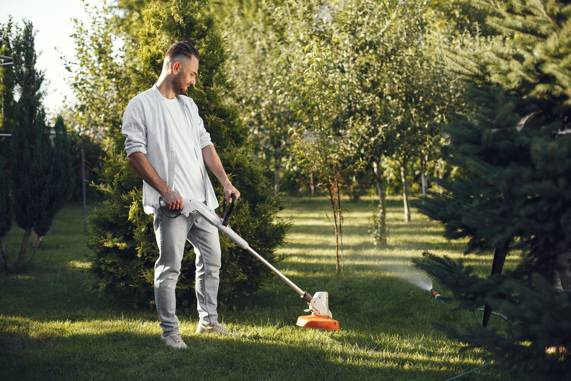 Man using a string trimmer to cut grass in a green, sunlit yard.