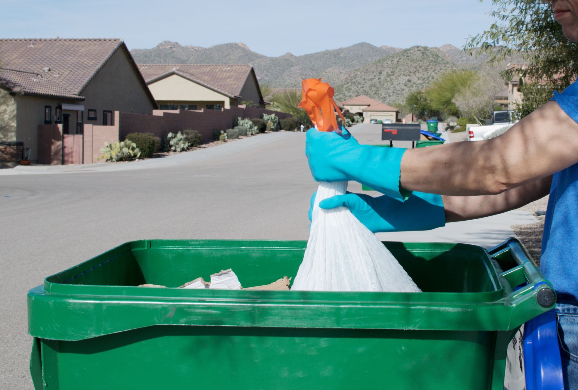 Person in blue gloves throwing a white trash bag into a green garbage bin on a suburban street.