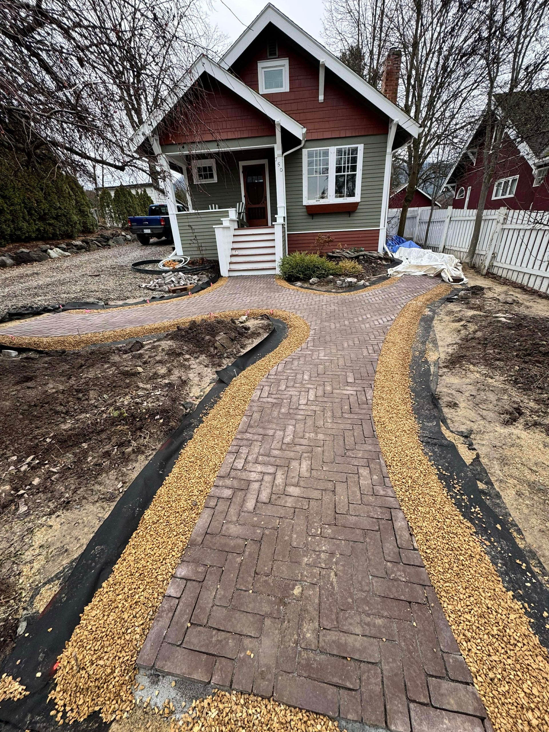 Brick pathway leading to a small house with red roof and green siding. Yellow gravel borders the path.