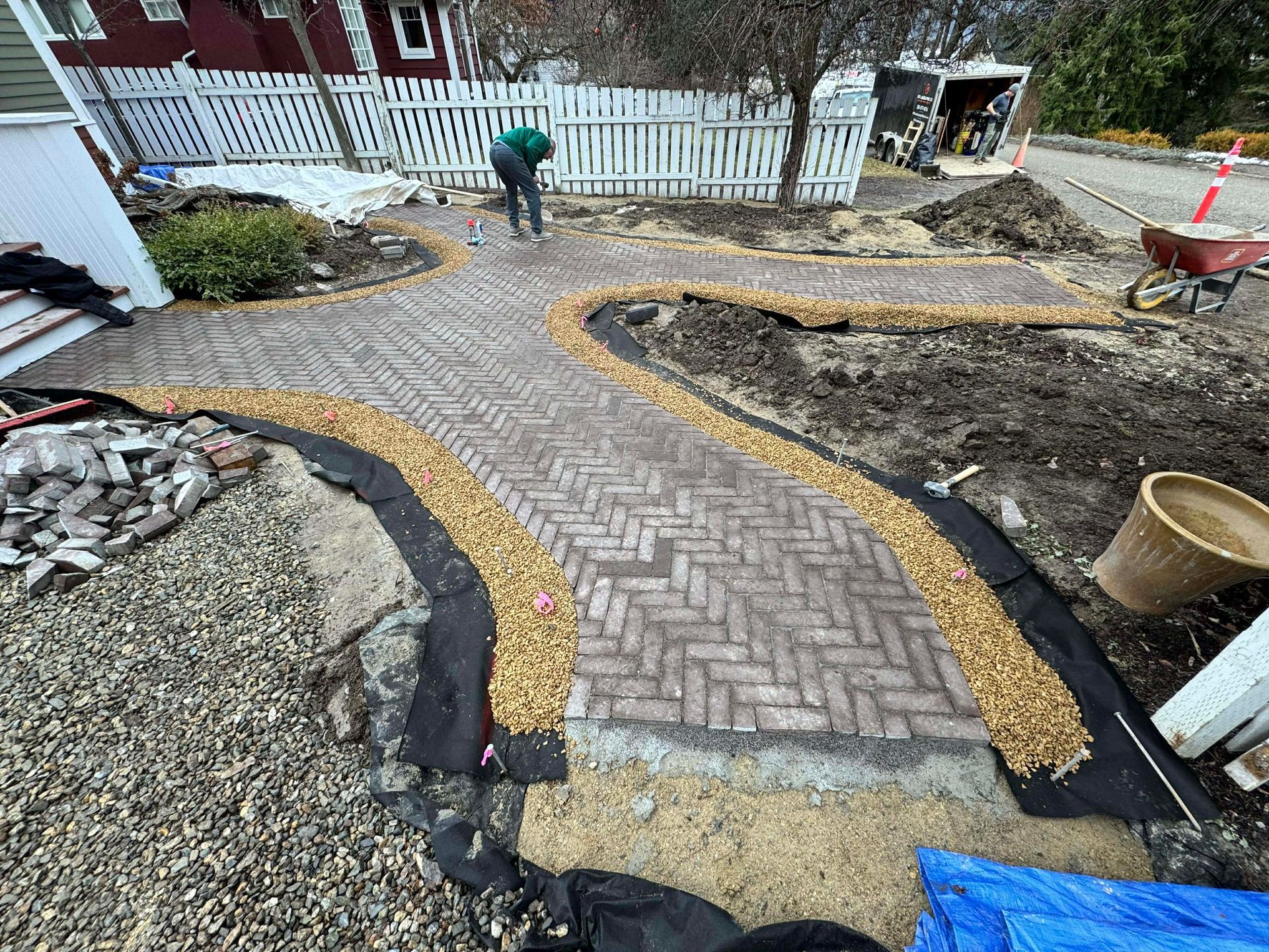 Brick pathway under construction with person working. Gravel border, dirt, and tools present.