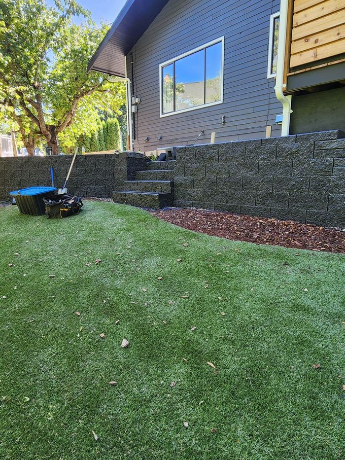 Backyard with artificial turf, stone wall, and blue lawnmower. House in the background.