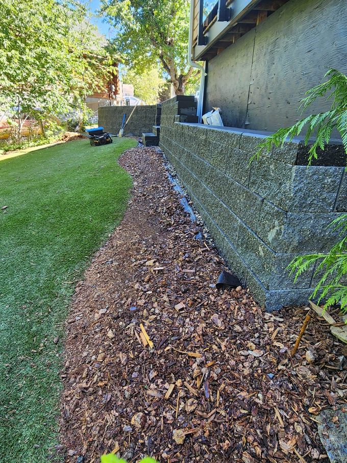 Mulched area along a textured concrete wall next to green grass and a building.