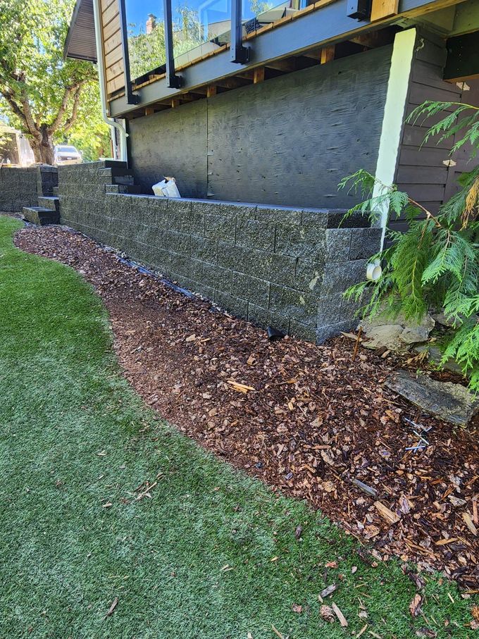 Stone retaining wall beside grass and mulch, under a house with a deck.