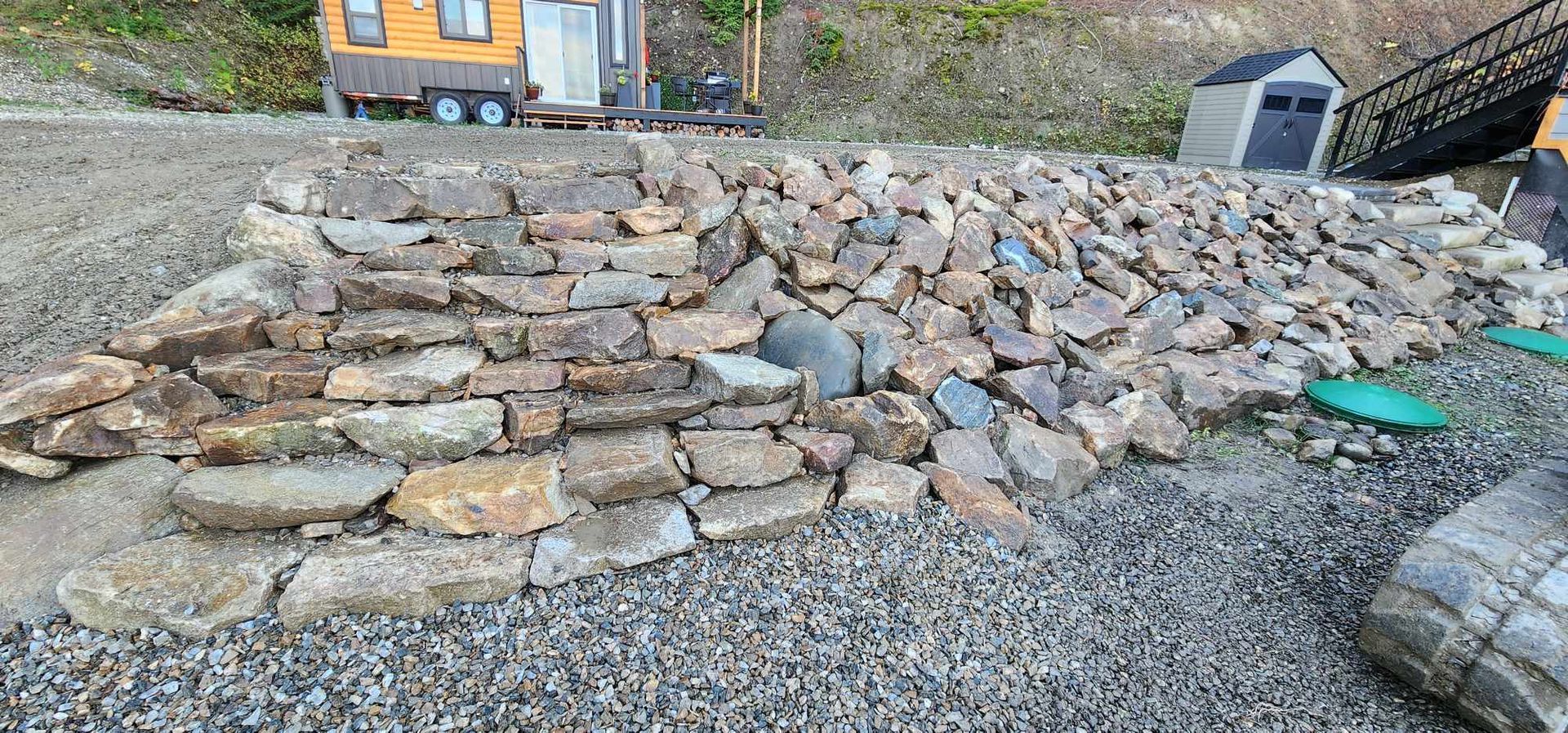 Stone steps leading up a sloped, gravelly area with a small shed and tiny house in the background.