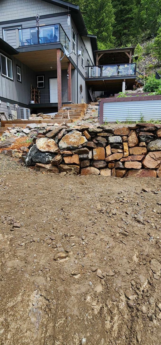 House on a sloped hillside with stone retaining wall and dirt in front.
