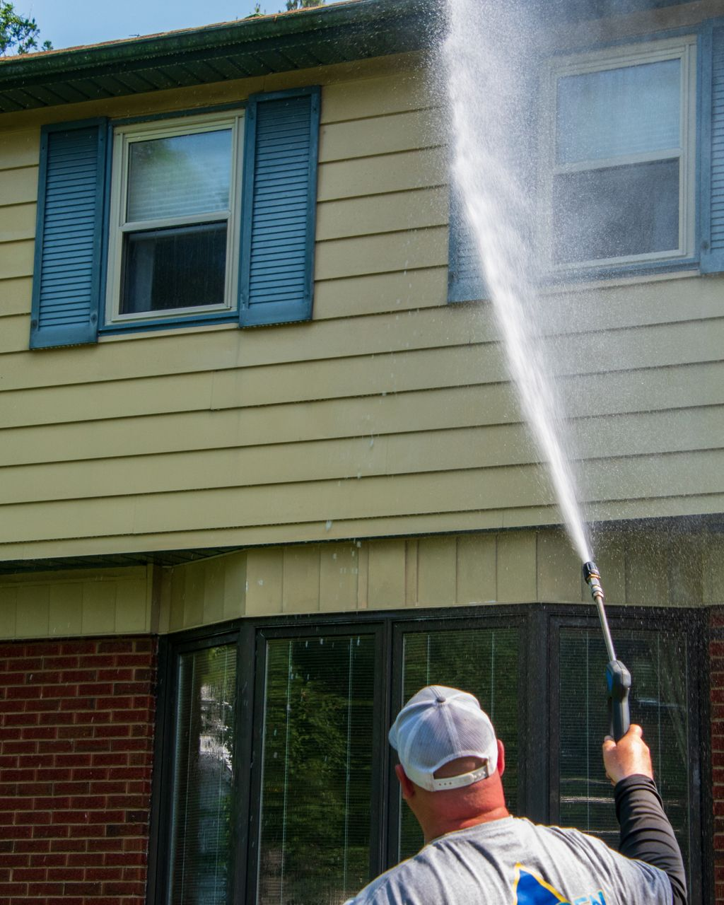 Person power washing the side of a two-story house with light blue shutters.