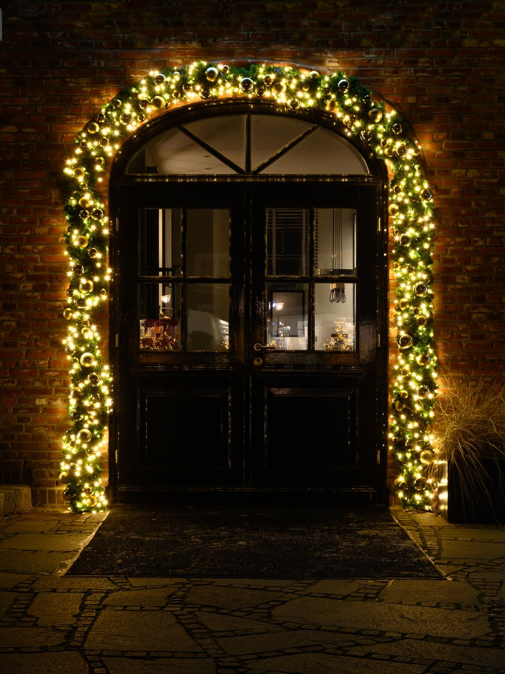 Black double doors with Christmas lights framing an arched entryway of a brick building.