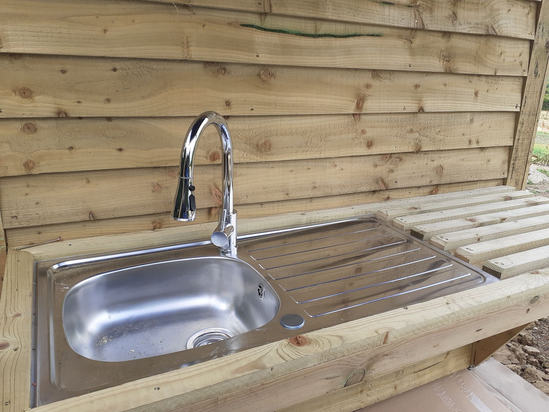 Stainless steel sink with a faucet set in a wooden structure outdoors.