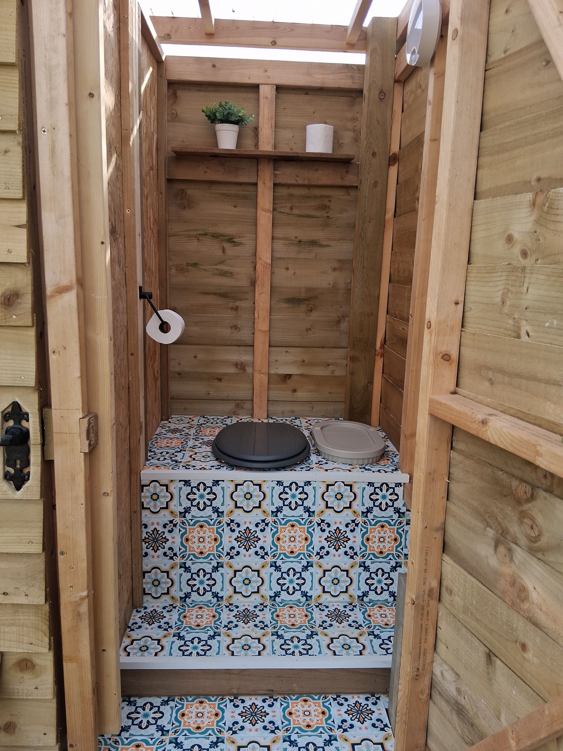 Wooden outhouse interior with patterned floor tiles, toilet, shelf with plants, and a roll of toilet paper.